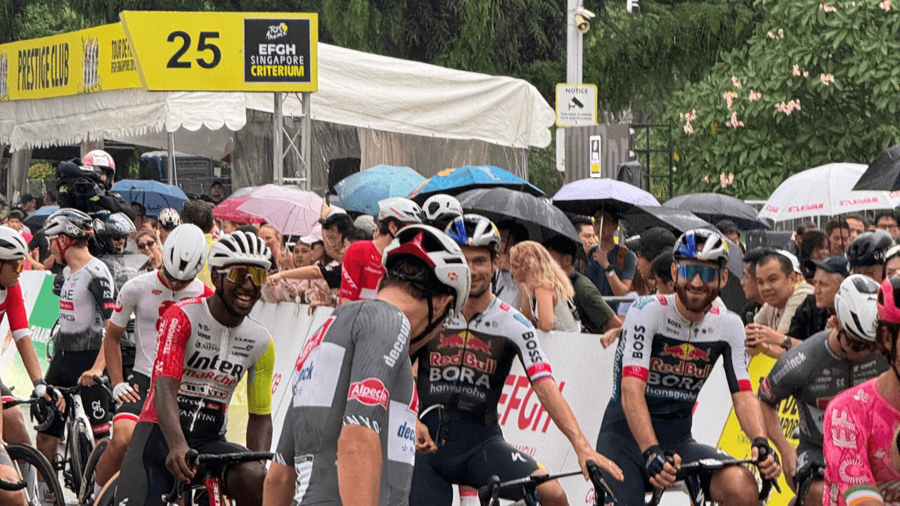 Professional cyclists smiling and chatting at the start line of the Tour de France EFGH Singapore Criterium under light rain, with spectators holding umbrellas in the background.